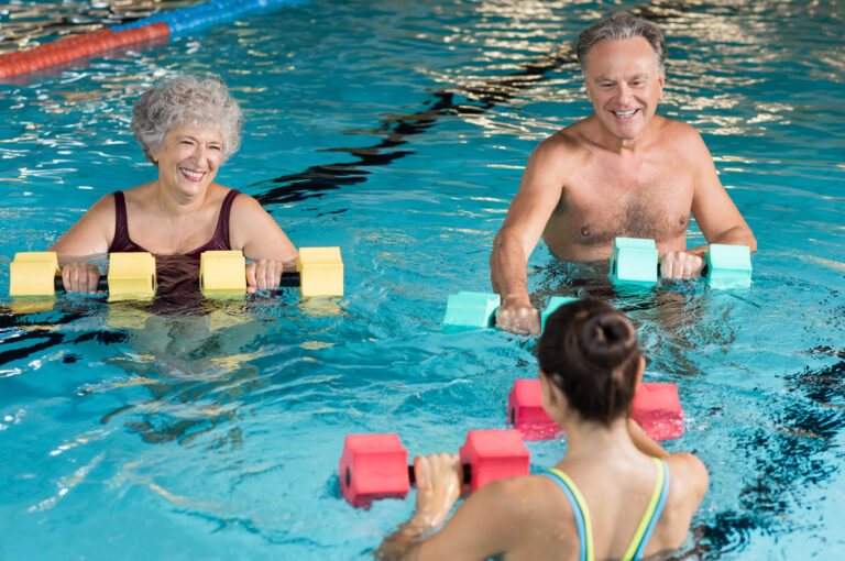 Senior couple in training session of aqua aerobics using dumbbells in swimming pool. Mature man and old woman practicing aqua fitness together. Healthy and fit senior couple enjoying their retirement in aqua aerobics training.