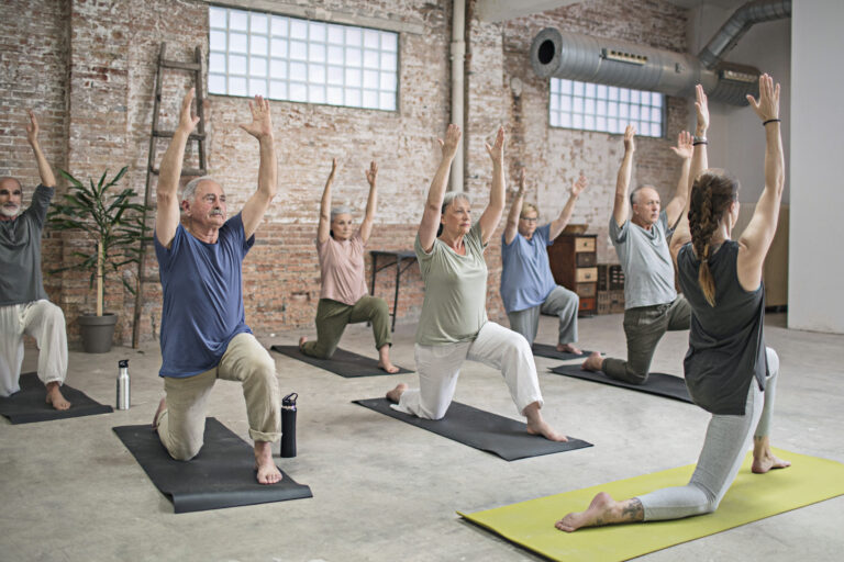 Senior people and fitness instructor exercising in warrior position at gym. Active seniors are exercising on exercise mats at health club. They are performing yoga together in studio.